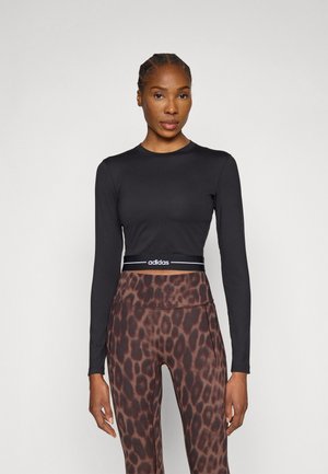 Bluză neagră cu mâneci lungi, cu un design ajustat și logo-ul Adidas pe talie, combinată cu leggings maro cu imprimeu leopard.