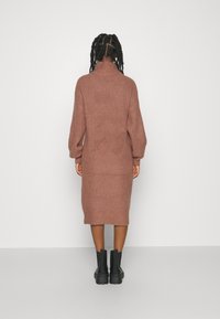 Robe tricotée rose avec un col montant, des manches longues et des poignets côtelés. Longueur mi-mollet avec une coupe décontractée, portée avec des bottines noires.