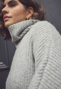 Maglione a collo alto grigio chiaro con texture a costine. Presenta un colletto alto e una vestibilità comoda, evidenziando un morbido materiale a maglia.