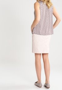 Kvinde iført ærmeløs bluse i taupe med prikker, lys rosa knælang nederdel og beige flade sko, stående med hænderne i lommerne, vendt væk.