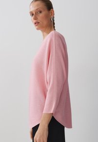 Pull en maille côtelée rose clair, avec une coupe décontractée, des manches trois-quarts et un décolleté arrondi. Il présente un ourlet asymétrique et une texture subtile.