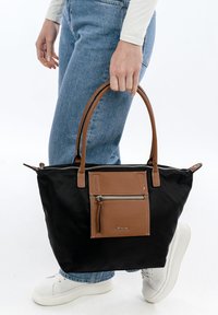 Borsa a tracolla in nylon nero con dettagli in pelle marrone, dotata di una tasca frontale con zip e due manici. Indossata con jeans azzurri chiari e scarpe da ginnastica bianche.