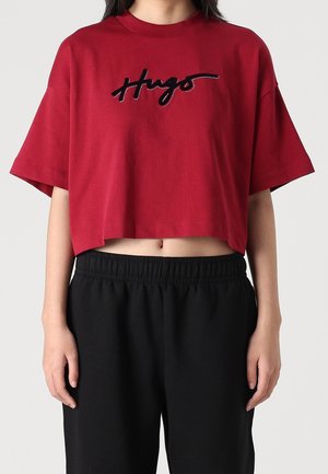 Nainen, joka pukeutunut punaiseen lyhytmidalliseen T-paitaan, jossa rinnan kohdalla lukee "Hugs", ja mustiin joustavaan vyötäröhousuihin yksiväristä taustaa vasten.