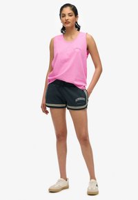 Camiseta sin mangas rosa con escote redondo y logo de "Superdry"; pantalones cortos azul marino con detalles blancos y cordón. Zapatillas blancas.