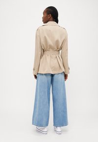 Beige belted trenchcoat med strukturerte skuldre, lagdelt bakside og knappe mansjetter, sammen med lyseblå vide jeans og hvite sneakers.