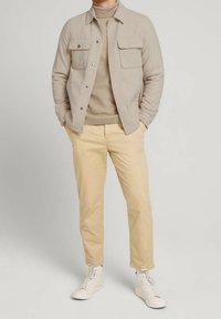 Giacca beige con bottoni e due tasche sul petto, dolcevita color cammello e pantaloni chino beige chiaro. Indossati con sneaker bianche alte.