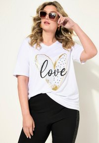 Bílý tričko s krátkým rukávem, které má zlaté a černé srdce a nápis "láska"; zkombinované s černými kalhotami s texturovaným bočním detailem.