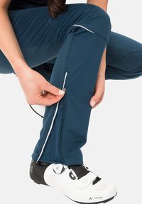 Blaue technische Hosen mit weißen Akzenten, ausgestattet mit einem Reißverschluss an den Knöcheln. Tragen mit einem weißen Radschuh, in einer hockenden Position gezeigt.