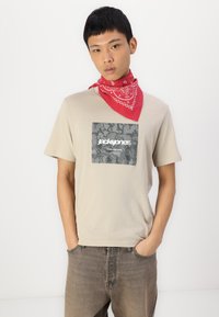 T-shirt beige à manches courtes avec un écusson en camouflage gris. Porté avec un jean en denim gris et un bandana rouge autour du cou.