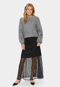 Szürke kötött pulóver gyöngy részletekkel, fekete csipkebabásszal párosítva. Az öltözet laza szabású és textúrált rétegekből áll.