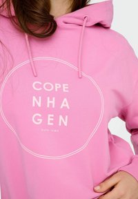 Rosa hoodie i bomull med en stor cirkulär vit grafik. Texten "COPENHAGEN" och koordinater tryckta i vitt framtill.