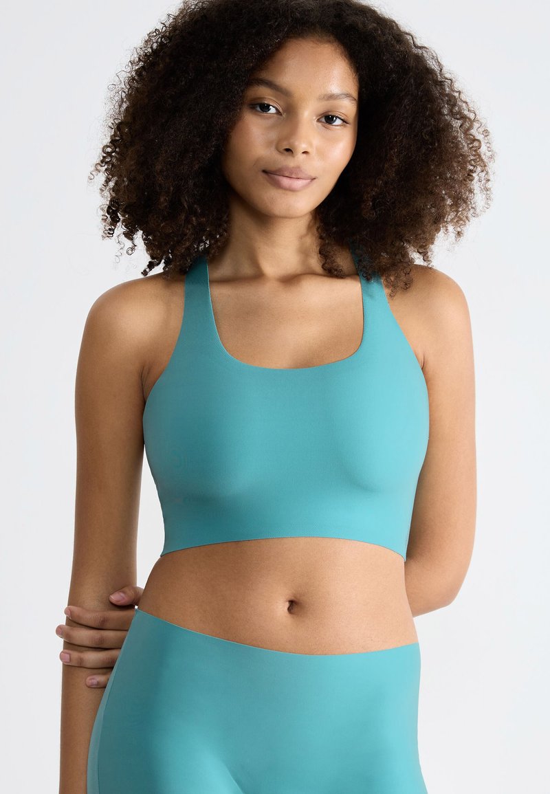 Turkis sportsbh med firkantet halsudskæring og brede stropper, lavet af glat, strækbart materiale. Har et minimalistisk design og en sømløs udseende.