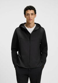 Svart zip-up hoodie med en slät textur, med en framdragkedja, två sidofickor och en passformshuva. Bärs över en vit skjorta.