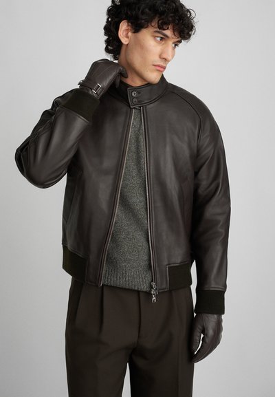 Veste bombardier en cuir marron avec des poignets et un col côtelés verts, dotée d'une fermeture éclair à l'avant et de poches latérales, portée sur un pull gris.
