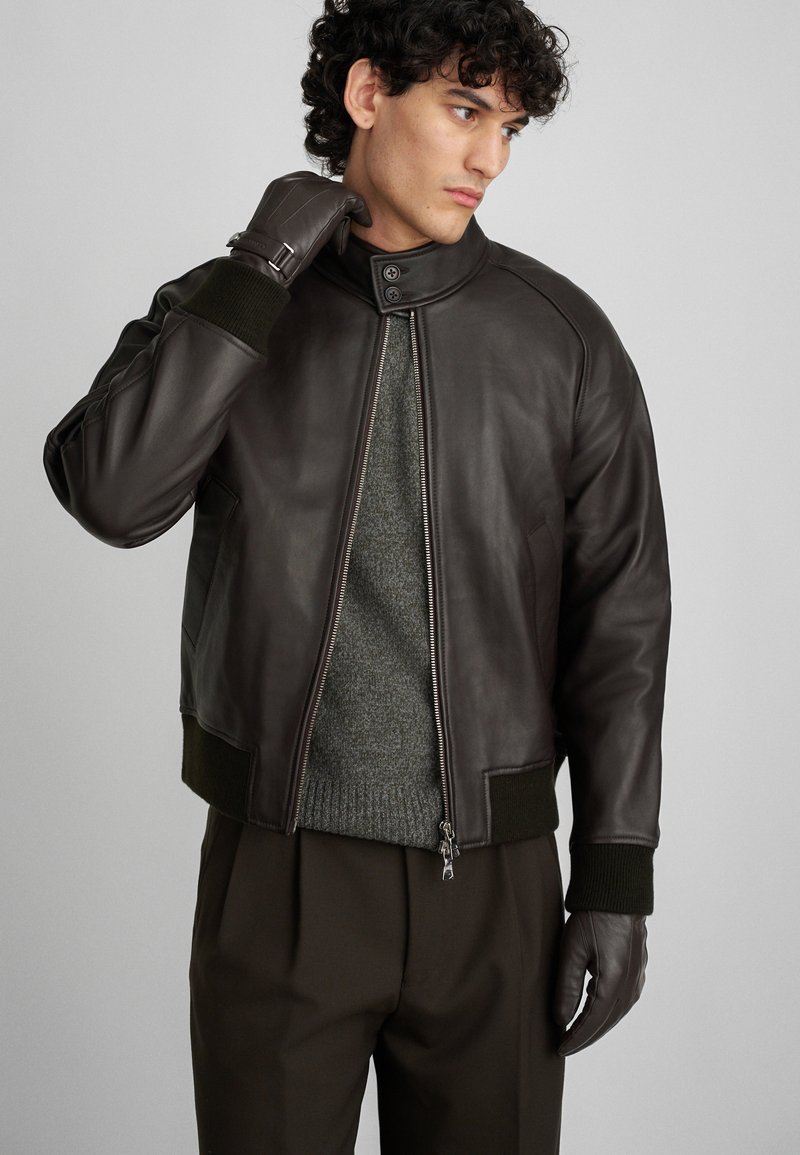 Καφέ δερμάτινο μπουφάν βόμβας με ρ ιμπ πράσινα μανίκια και γιακά, με φερμουάρ μπροστά και πλαϊνές τσέπες, φορεμένο πάνω από ένα γκρι πουλόβερ.