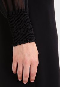 Main à peau claire portant une manche longue noire avec un poignet élastiqué froncé et un bord à volants sur un fond noir.