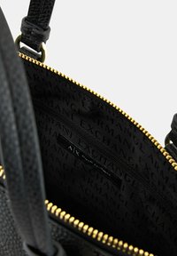 Interno di una borsa nera texturizzata con cerniera d'oro, dotato di una tasca con zip e rivestimento in tessuto marchiato con il testo "Armani Exchange".
