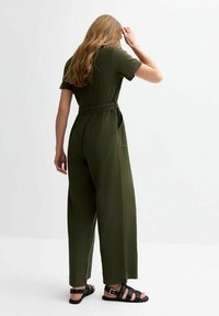 Khaki green jumpsuit with short sleeves, loose fit, a tied waist belt, side pockets, and back detail visible. Sandals with black straps.