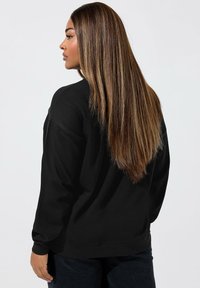 Felpa nera con vestibilità rilassata, polsini e orlo a costine. Texture morbida, indossata con pantaloni scuri, che mettono in mostra capelli lunghi e lisci.