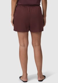 Pantaloni corti color borgogna con vita elasticizzata, tessuto liscio e vestibilità comoda, che terminano appena sopra la coscia. Indossati con scarpe nere senza lacci.