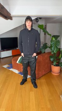 Noor inimene mustas kapuutsiga pusa ja pükstega seisab käed taskutes moodsas elutoas oranži diivani ja suure potitaimedega.