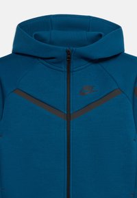 Fato de treino azul da Nike com fecho, detalhes diagonais pretos e colar alto, mostrando a parte superior do torso e o capuz.