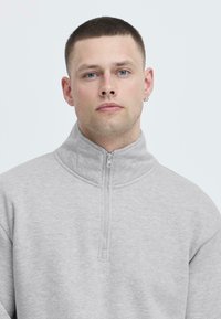 Lys grå kvart-zip genser laget av mykt stoff. Høy krage, stikkingdetaljer, og en jevn tekstur med minimal mønstring.