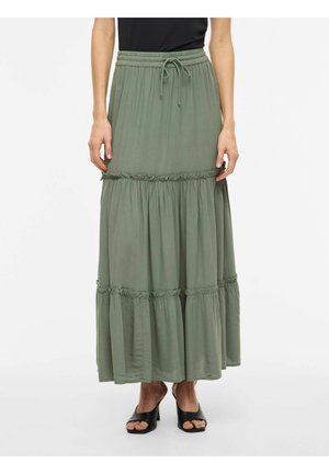 Kvinne iført olivengrønn lagdelt maxiskjørt med elastisk midje og svarte åpne heeled sandaler, stående på hvit bakgrunn.