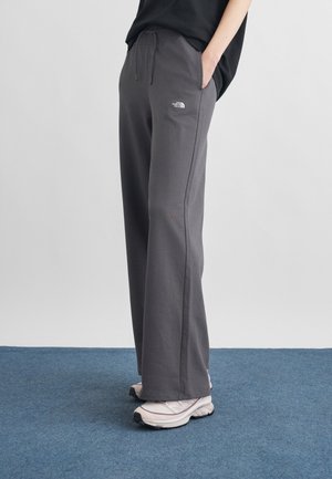 Άτομο που φοράει σκουρόχρωμο γκρι παντελόνι North Face και λευκά αθλητικά παπούτσια, στέκεται σε μπλε χαλί με τα χέρια στις τσέπες.