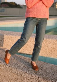 Bailarinas de cuero marrón, jeans azules con dobladillo desgastado y un suéter rosa suave. De pie sobre una superficie de concreto texturizado cerca del agua.