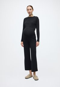 Macacão de maternidade preto com rachas, mangas compridas e corte descontraído, apresentando um design de perna larga e bolsos laterais. Calçado casual completa o visual.