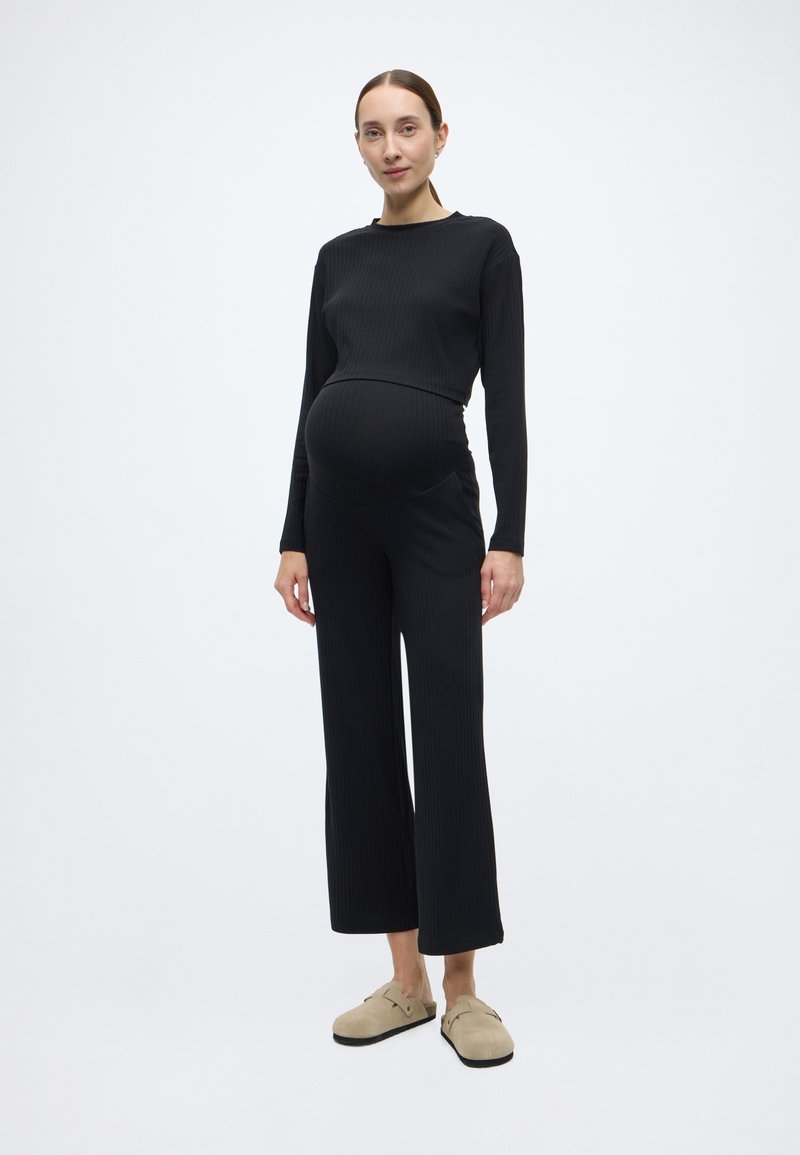 Macacão de maternidade preto com rachas, mangas compridas e corte descontraído, apresentando um design de perna larga e bolsos laterais. Calçado casual completa o visual.