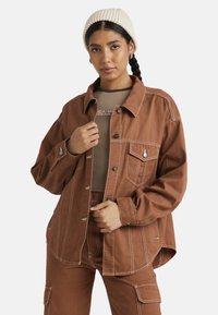 Giacca oversize marrone con cuciture bianche, chiusura a bottoni e tasca sul petto. Indossata sopra un top beige, abbinata a pantaloni beige e un berretto crema.