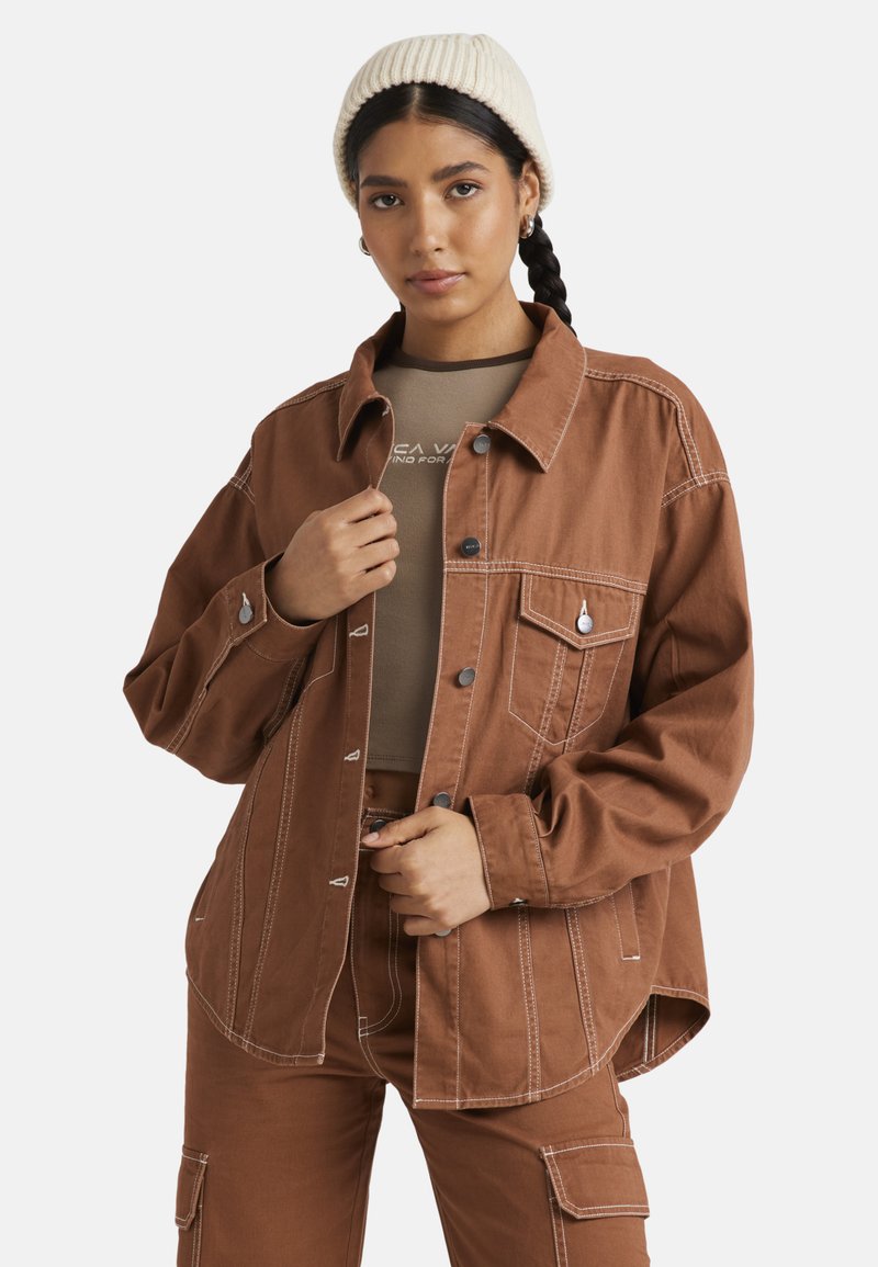 Giacca oversize marrone con cuciture bianche, chiusura a bottoni e tasca sul petto. Indossata sopra un top beige, abbinata a pantaloni beige e un berretto crema.