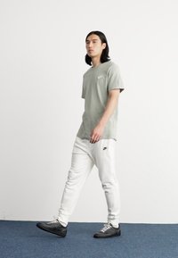 T-shirt de algodão verde claro com um logo no peito, combinado com calças brancas que apresentam um logo lateral. Ténis pretos completam o conjunto.