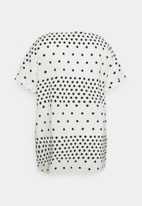 Blouse blanche à manches courtes avec des pois noirs disposés en bandes horizontales de densité variable.