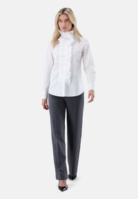White ruffled blouse with a high collar and front button closure, paired with gray trousers and black pointed-toe heels, featuring a smooth texture.