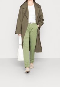Khaki trenchcoat, vit t-shirt, ljusgröna byxor, beige slip-ons, håller en vit väska; kännetecknas av avslappnad passform och enkel design.