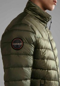Giacca puffer verde oliva con collo alto, texture trapuntata e una toppa rotonda nera e arancione sulla spalla sinistra. Chiusura a zip.