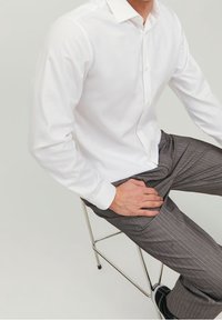 Camicia bianca a maniche lunghe con colletto diritto, abbinata a pantaloni grigi a righe sottili. Modello seduto su una sedia di metallo.