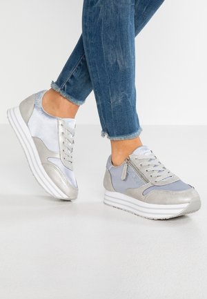 Stopy w srebrno-szarych sneakersach na platformie z bocznymi zamkami i sznurowadłami, w połączeniu z niebieskimi jeansami ze strzępionymi nogawkami na białym tle.