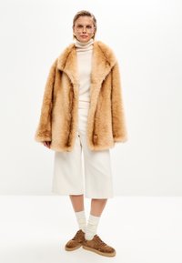 Tan faux fur-frakke med stor krave, båret over en cremefarvet turtleneck, parret med brede hvide bukser og brune hjemmesko i ruskind.