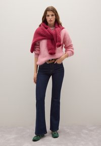 Pullover rosa in peluche con maniche lunghe, abbinato a jeans a palazzo scuri. Una sciarpa rossa avvolta attorno al collo e scarpe verdi completano il look.