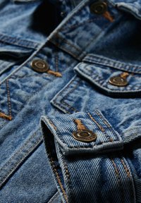 Jeansjacka i ljusblått med gyllene sömmar, med metallknappar och fickor med lock, som visar en texturerad tyg.