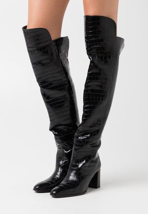 Bottes montantes noires en similicuir brillant à texture crocodile, avec un bout pointu effilé et un talon épais. Elles présentent une découpe allongée en haut.