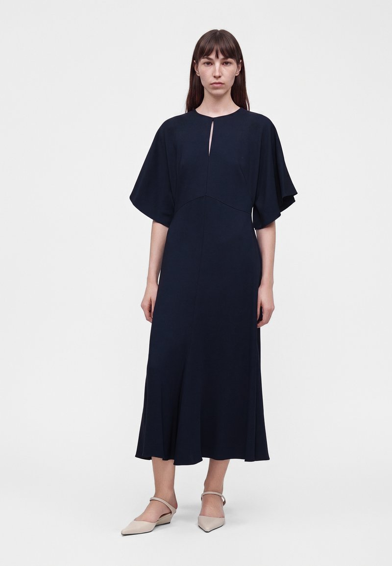 Femme portant une longue robe noire à manches larges avec une petite fente à l'avant, associée à des chaussures beiges à bout pointu avec des brides.