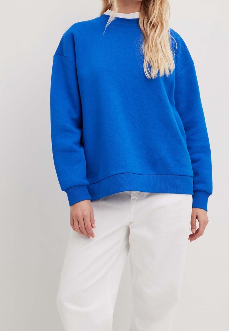 Sweat-shirt en coton bleu avec un col rond, des épaules tombantes et des poignets en côte. Assorti avec un pantalon blanc pour le contraste.