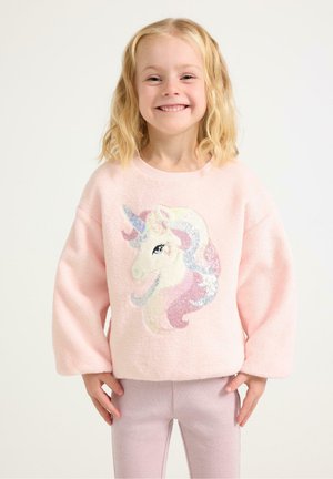 Hymyilevä nuori tyttö, jolla on vaaleat hiukset, pukeutuneena vaaleanpunaiseen villapaitaan, jossa on kimaltava yksisarvinen, ja vaaleanpunaisiin housuihin, seisomassa valkoista taustaa vasten.