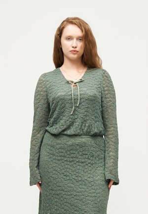 Jeune femme aux longs cheveux bruns portant une robe verte en dentelle à manches longues avec un col noué sur un fond clair uni.