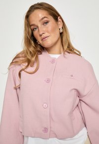 Svetloružová skrátená bunda s textúrovanou tkaninou, vybavená štyrmi zodpovedajúcimi gombíkmi a malou vreckom na hrudi. Mäkký a uvoľnený strih.